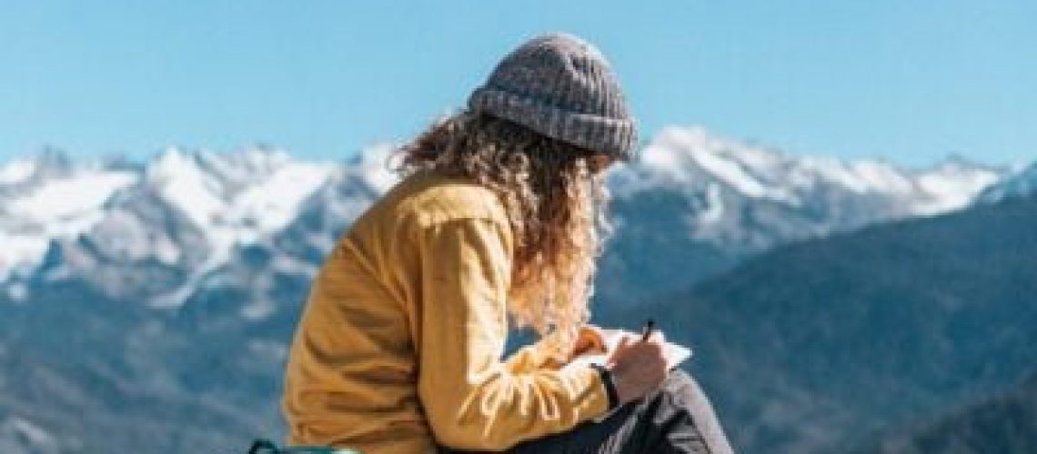 woman writing while sitting on hill near mountain